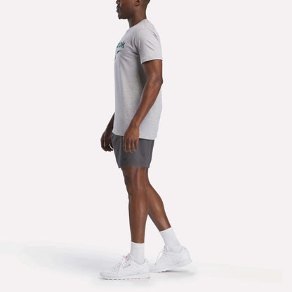 A man stands in profile against a plain background, wearing a gray Reebok Men's Stacked Vector Graphic T-Shirt, dark gray shorts, white socks, and white athletic shoes.