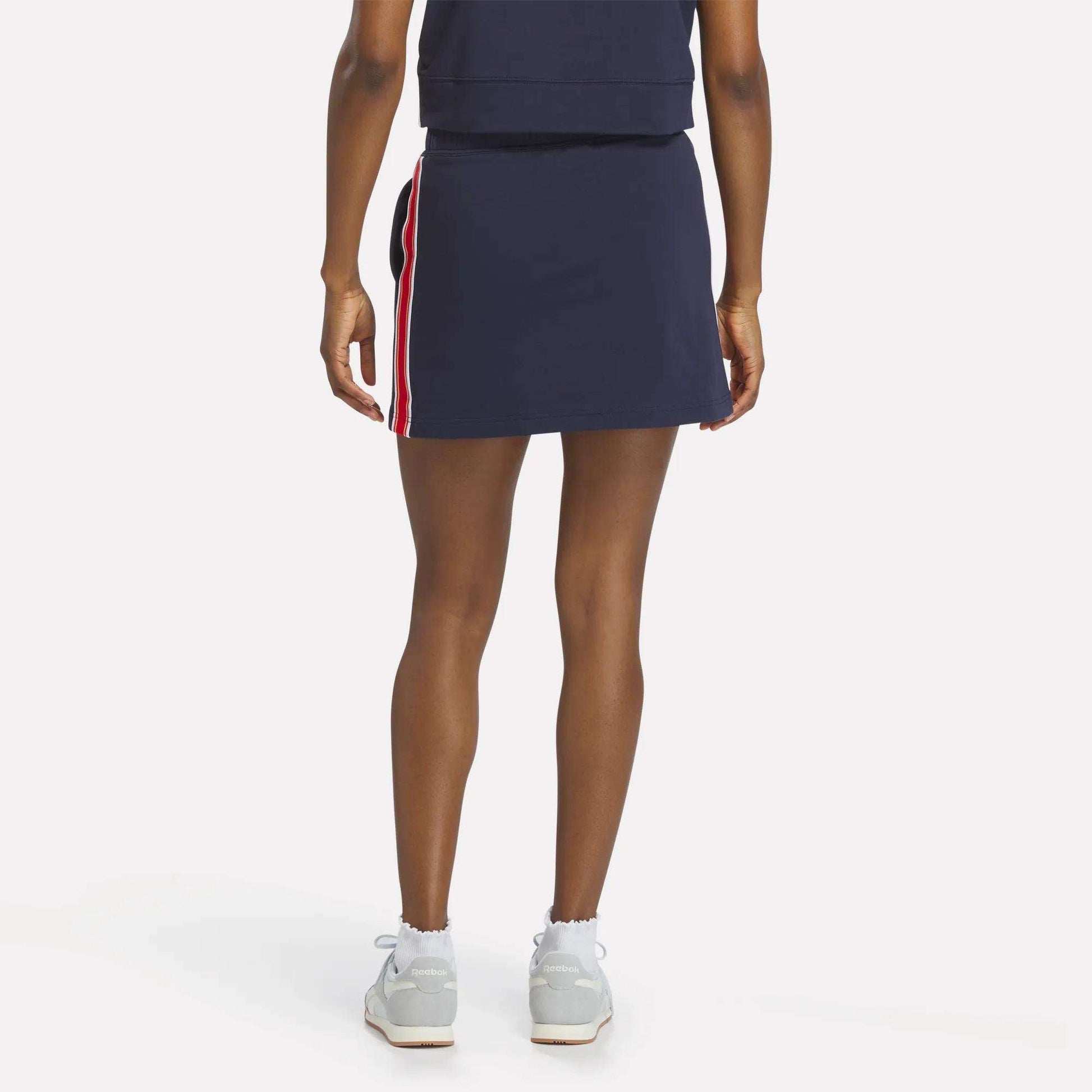A woman faces away, wearing the Reebok Women's Logo Skirt in navy with red and white side stripes, paired with a matching navy top, white socks, and light sneakers against a plain white background.
