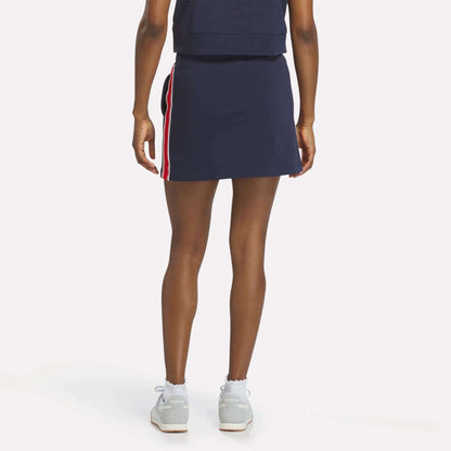 A woman faces away, wearing the Reebok Women's Logo Skirt in navy with red and white side stripes, paired with a matching navy top, white socks, and light sneakers against a plain white background.