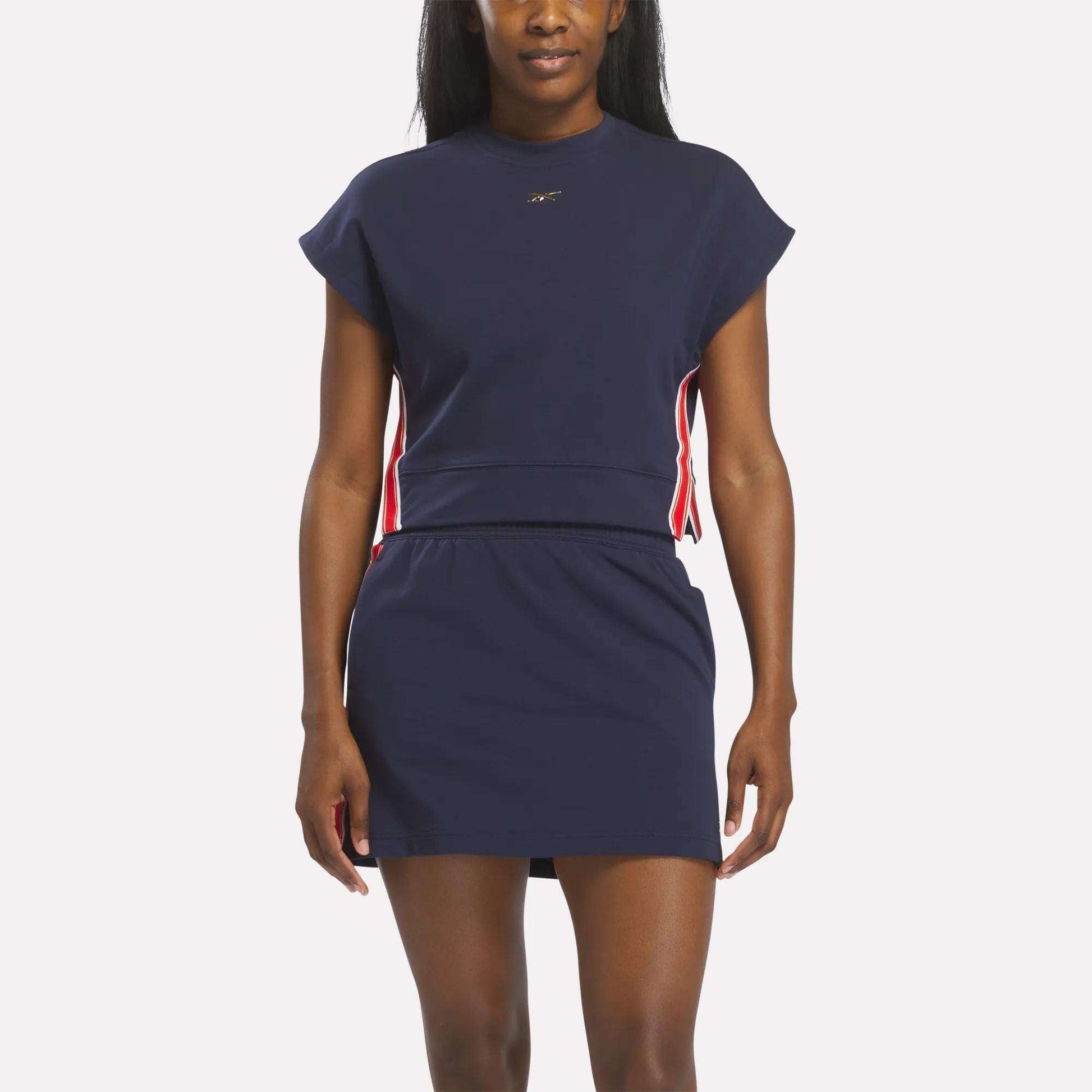 A woman wearing the Reebok Women's Logo Short Sleeve Crewneck Sweatshirt in Vector Navy stands against a white background. The fitted waist and sporty crewneck style give this casual top a fresh, athletic look.