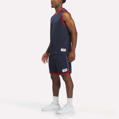 A man wears the Reebok Men's United Jersey Tank in navy with matching red-trimmed shorts, paired with white socks and sneakers, standing against a plain white background.