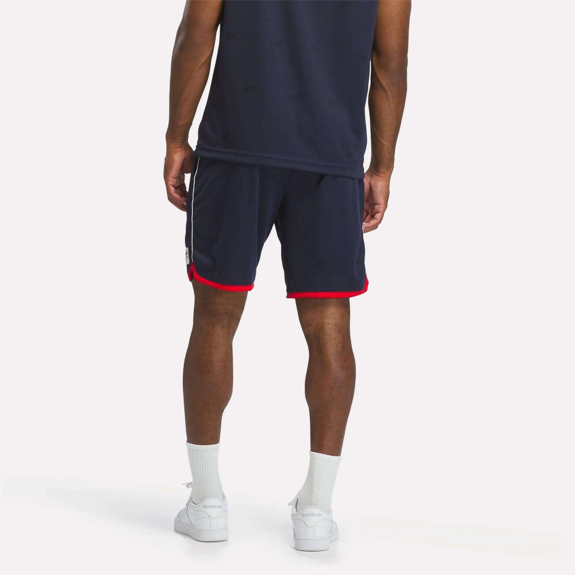 A person wearing Reebok Men's United Shorts 9" in navy with red trim, a matching shirt, white socks, and white sneakers stands with their back to the camera against a plain background.