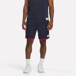 A person in a navy athletic outfit with a sleeveless top and Reebok Men's United Shorts 9" in Vector Navy, plus white socks and sneakers, stands against a plain light background.