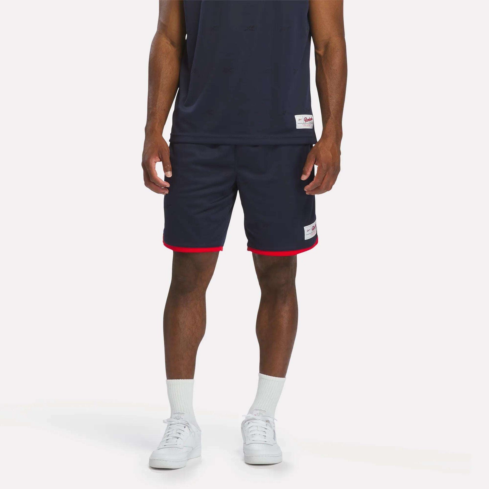 A person in a navy athletic outfit with a sleeveless top and Reebok Men's United Shorts 9" in Vector Navy, plus white socks and sneakers, stands against a plain light background.