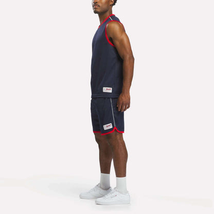 A man stands sideways wearing Reebok Men's United Shorts 9" in navy with red trim, paired with a sleeveless athletic top, white socks, and white sneakers against a plain white background.