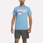 A man stands facing forward against a plain white background, wearing a light blue heather Reebok Men's Stacked Vector Graphic T-Shirt and dark gray athletic shorts.