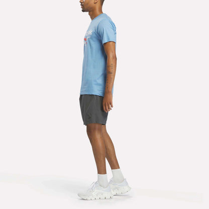A person stands in profile facing left, wearing a light blue Reebok Men's Stacked Vector Graphic T-Shirt, dark athletic shorts, white socks, and white Reebok sneakers against a plain white background.