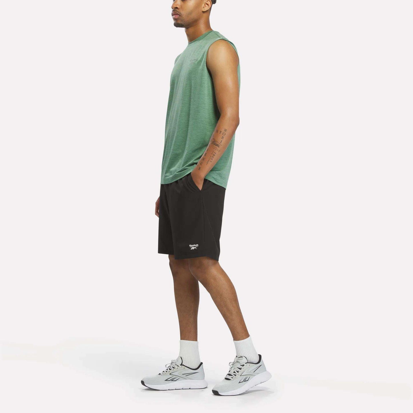 A person stands sideways against a plain white background, wearing the Reebok Men's Elite Sleeveless Top in green—crafted from cotton flex knit—paired with black shorts, white crew socks, and white Reebok athletic shoes.