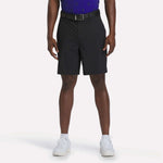 A person stands against a plain white background wearing a blue shirt and the Reebok Men's Pull-On Golf Shorts 8.5" in black, which feature four-way stretch for added comfort. The look is completed with white sneakers.