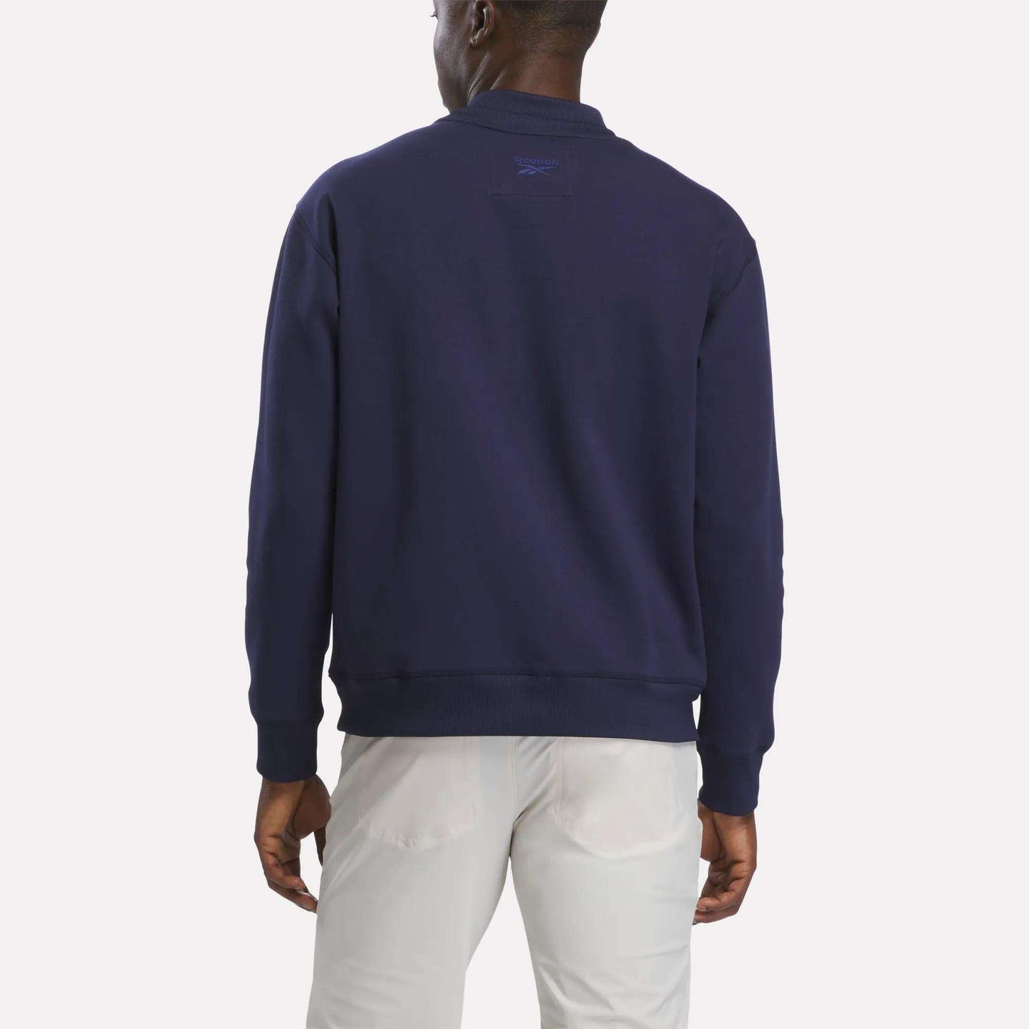 A man stands facing away, wearing a men's Reebok Signature Golf Crewneck Sweatshirt in navy blue featuring RBK-DRY technology, paired with light-colored pants against a plain white background.