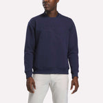 A man stands against a plain white background wearing the Reebok Men's Signature Golf Crewneck Sweatshirt in Midnight Blue, which features embossed lettering and advanced RBK-DRY technology, paired with white pants.