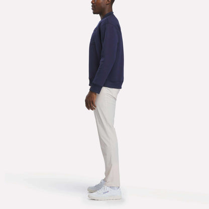 A person stands sideways against a plain white background, wearing the Reebok Men's Signature Golf Crewneck Sweatshirt in navy, paired with light-colored pants and white sneakers.