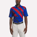 A man wearing a Reebok Men's Tournament Polo Shirt in Vector Blue, Navy, and Red stands with his arms at his sides against a plain background, paired with light-colored pants.