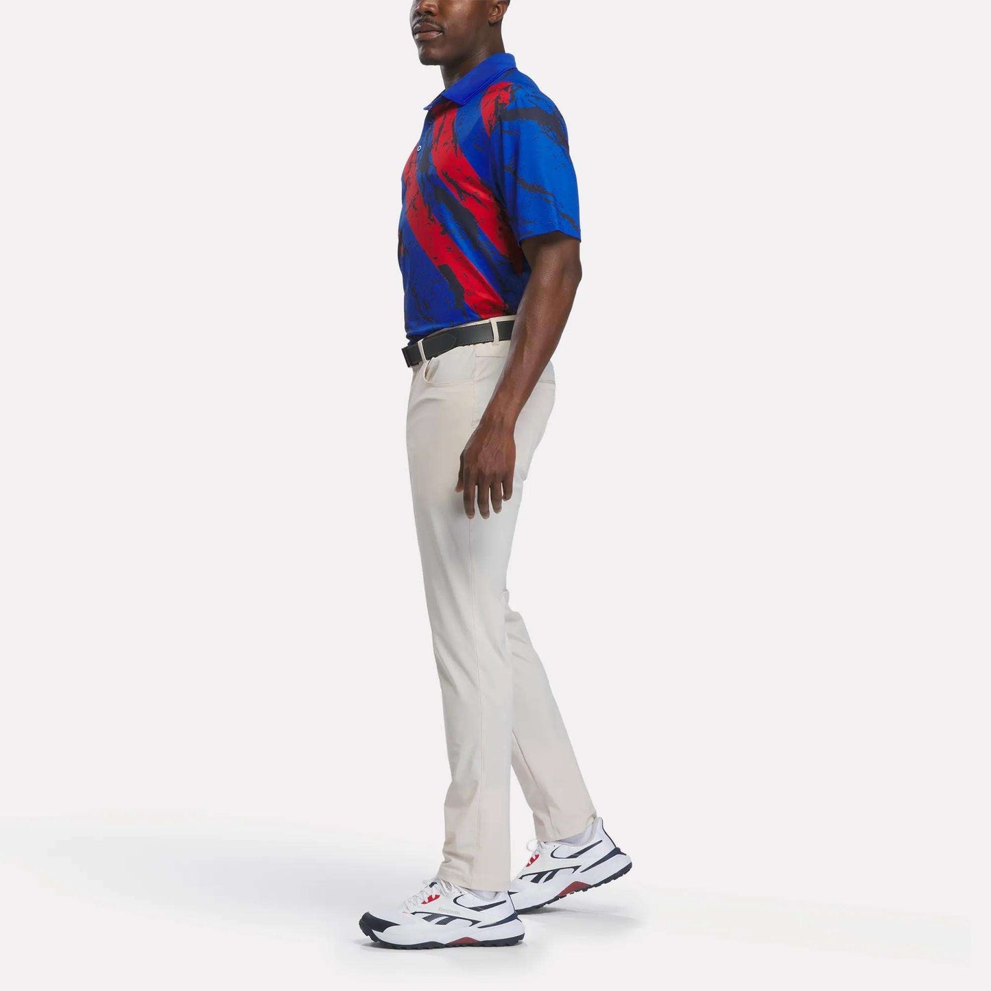 A man stands sideways wearing a Reebok Men's Tournament Polo Shirt in blue and red, paired with beige pants, a black belt, and white athletic shoes featuring black and red accents, set against a plain white background.
