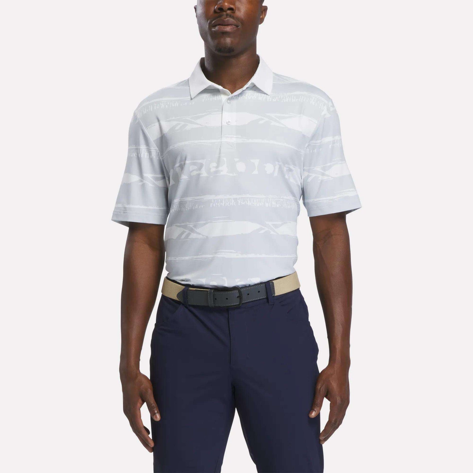 A man wears the Reebok Men's Tournament Polo Shirt in Grey/White, styled with navy blue pants and a beige belt, standing against a plain white background.