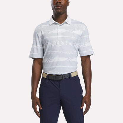 A man wears the Reebok Men's Tournament Polo Shirt in Grey/White, styled with navy blue pants and a beige belt, standing against a plain white background.