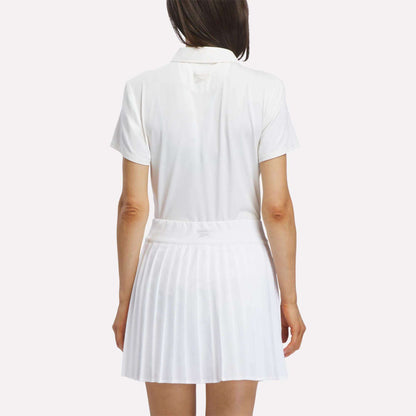 A woman stands facing away against a plain background, wearing a short-sleeved Reebok Women's Solid Core Polo Shirt in white and a white pleated skirt.