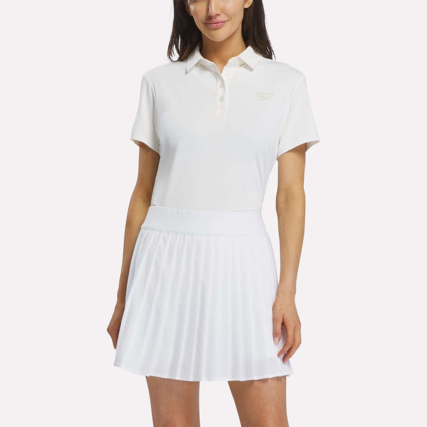A woman wearing a Reebok Women's Solid Core Polo Shirt in Chalk with a white pleated skirt, standing with one hand by her side and the other resting lightly on her skirt.