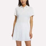 A woman wearing a Reebok Women's Solid Core Polo Shirt in Chalk with a white pleated skirt, standing with one hand by her side and the other resting lightly on her skirt.