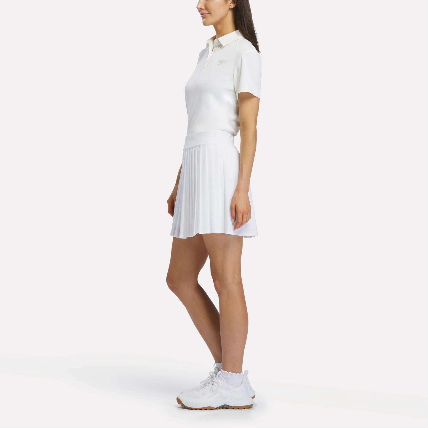 A woman stands sideways wearing the Reebok Women's Solid Core Polo Shirt in white, paired with a white pleated tennis skirt and white sneakers against a plain white background.