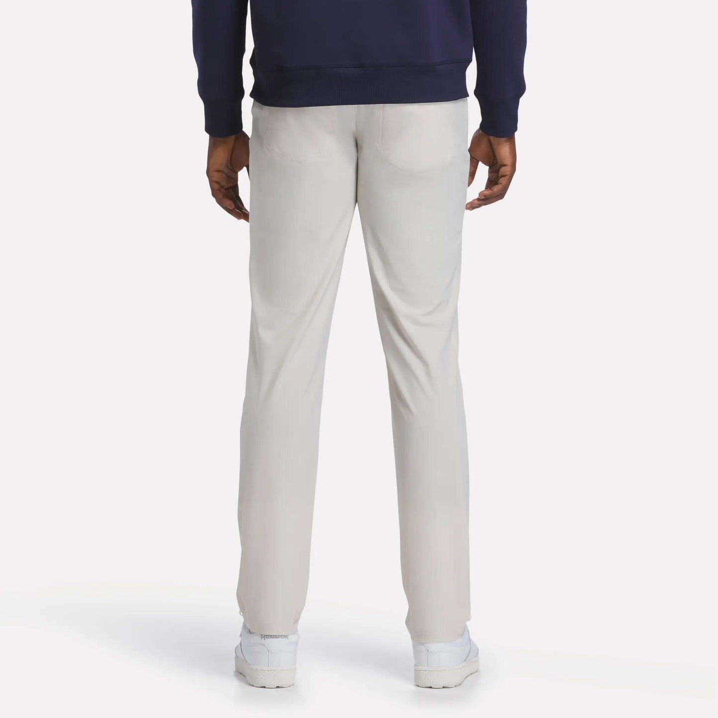 A person is standing facing away from the camera against a plain, light background, wearing Reebok Men's Favorite 5-Pocket Relaxed Fit Golf Pants in a light color, paired with a navy blue sweatshirt and white shoes.