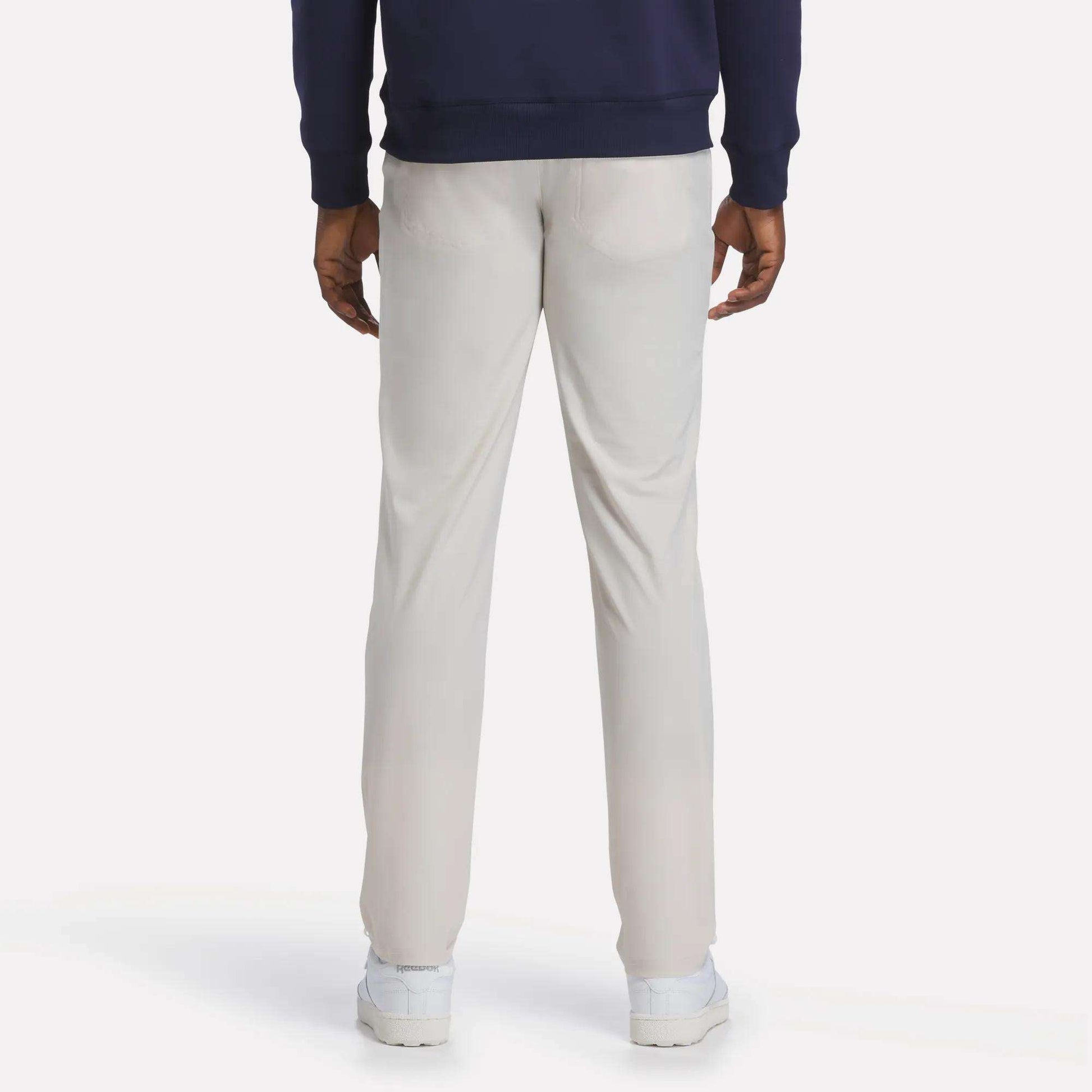A person is standing facing away from the camera against a plain, light background, wearing Reebok Men's Favorite 5-Pocket Relaxed Fit Golf Pants in a light color, paired with a navy blue sweatshirt and white shoes.