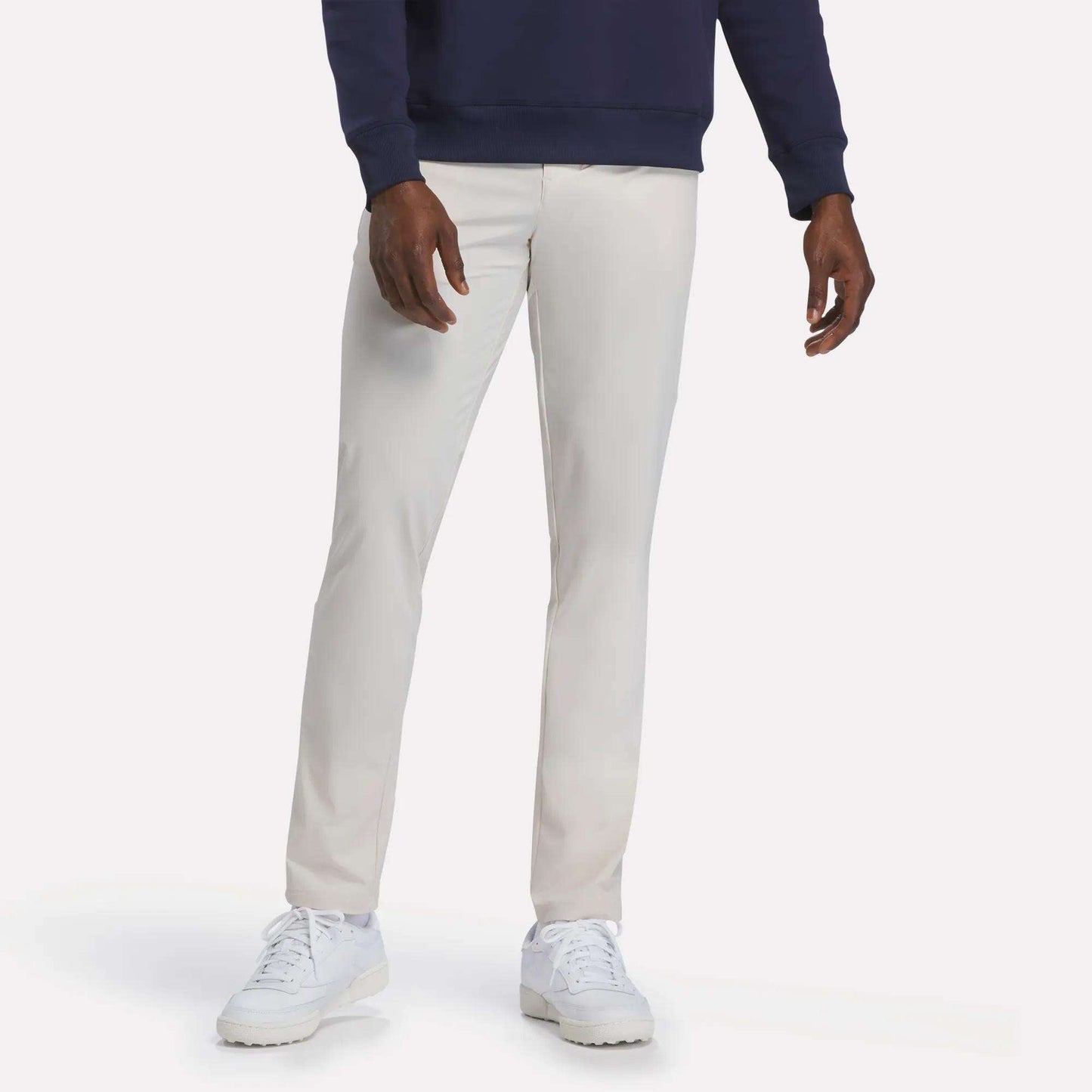 A person wearing Reebok’s Men's Favorite 5-Pocket Relaxed Fit Golf Pants in Moon Beam, a navy sweatshirt, and white sneakers stands against a plain white background. The wrinkle-resistant fabric of the pants ensures a sharp, polished look all day.