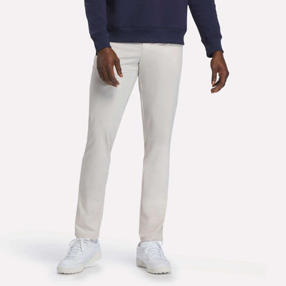 A person wearing Reebok’s Men's Favorite 5-Pocket Relaxed Fit Golf Pants in Moon Beam, a navy sweatshirt, and white sneakers stands against a plain white background. The wrinkle-resistant fabric of the pants ensures a sharp, polished look all day.
