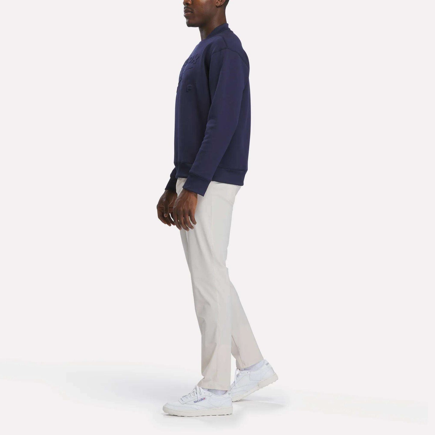 A person stands sideways against a plain white background wearing a navy sweatshirt, white sneakers, and Reebok Men's Favorite 5-Pocket Relaxed Fit Golf Pants in a wrinkle-resistant fabric.