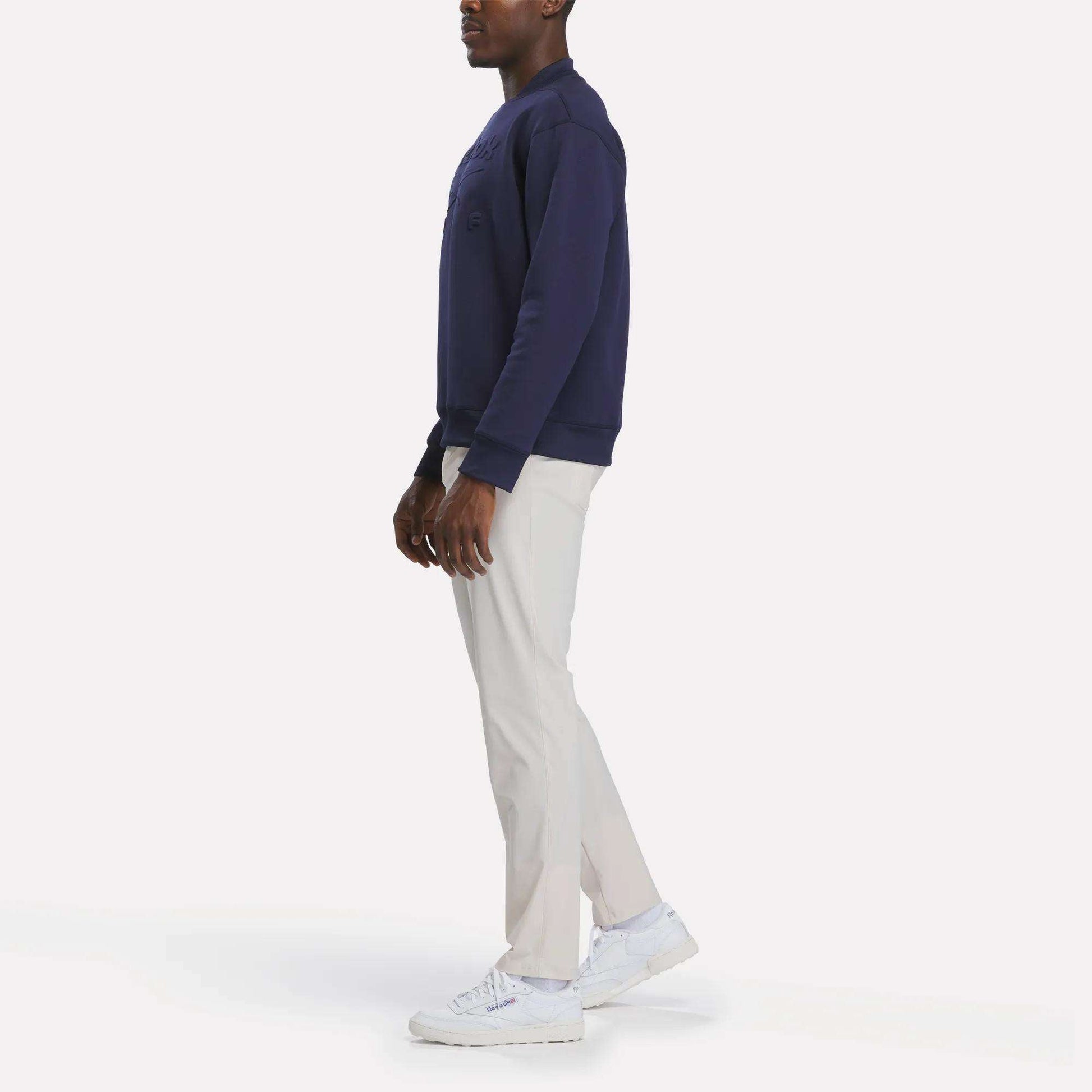 A person stands sideways against a plain white background wearing a navy sweatshirt, white sneakers, and Reebok Men's Favorite 5-Pocket Relaxed Fit Golf Pants in a wrinkle-resistant fabric.