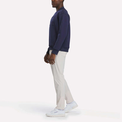 A person stands sideways against a plain white background wearing a navy sweatshirt, white sneakers, and Reebok Men's Favorite 5-Pocket Relaxed Fit Golf Pants in a wrinkle-resistant fabric.