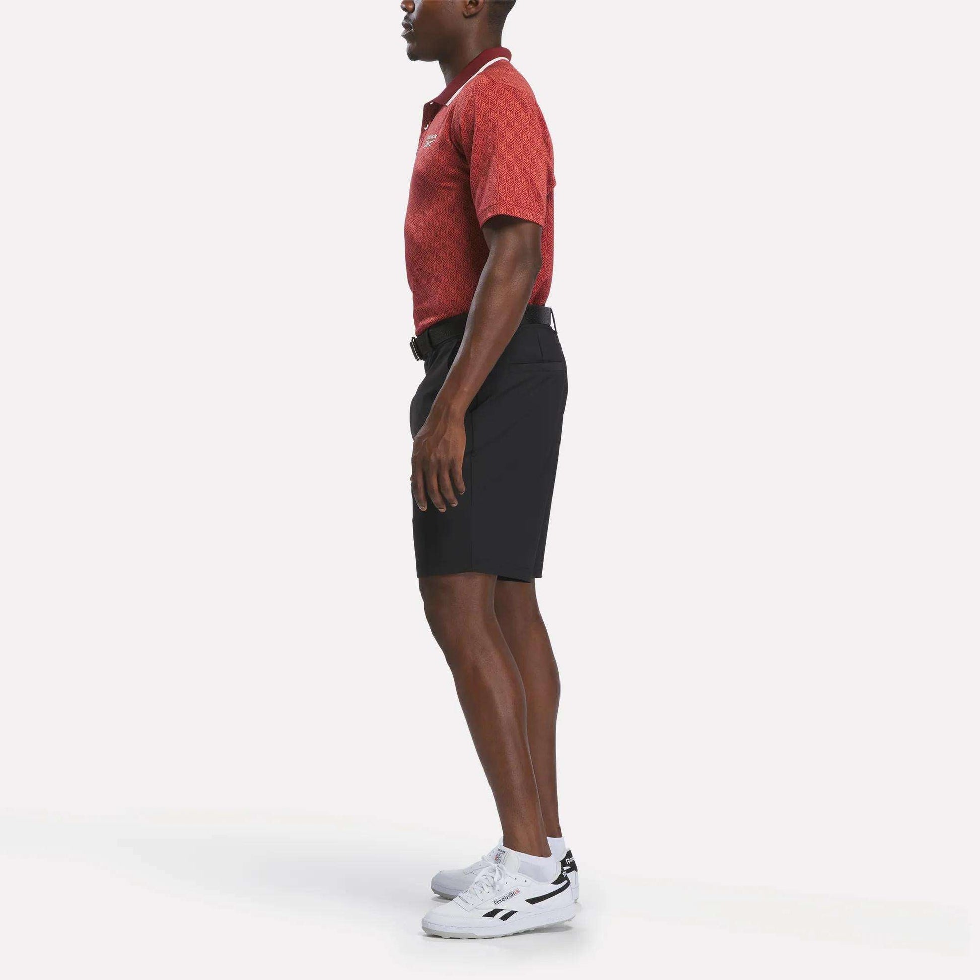 A man stands in profile wearing a Reebok Men's Jacquard Polo Shirt in red, paired with black shorts, white sneakers, and a black belt against a plain white background.