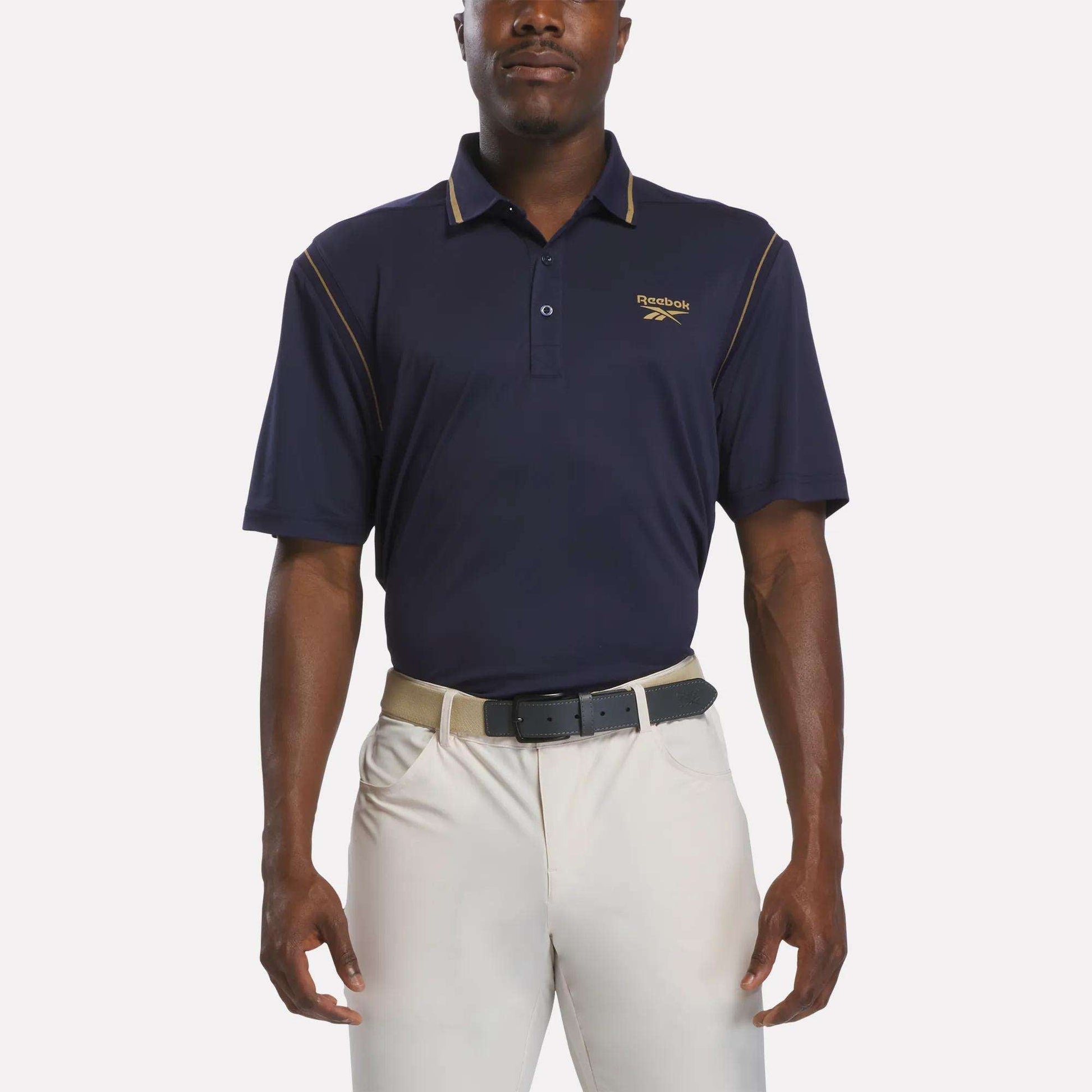 A man wearing a Reebok Men's Varsity Polo Shirt in Vector Navy with Trophy Gold piping, paired with light beige pants and a black belt, stands against a plain white background.
