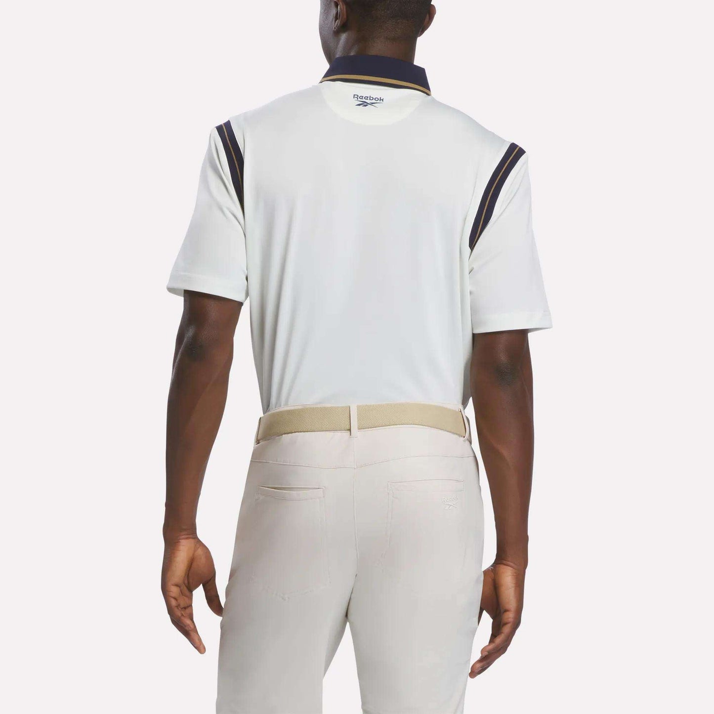A man is shown from the back wearing a Men's Varsity Polo Shirt by Reebok, featuring RBK-DRY fabric with navy accents. He pairs it with light beige pants and a light-colored belt. The background is plain white.