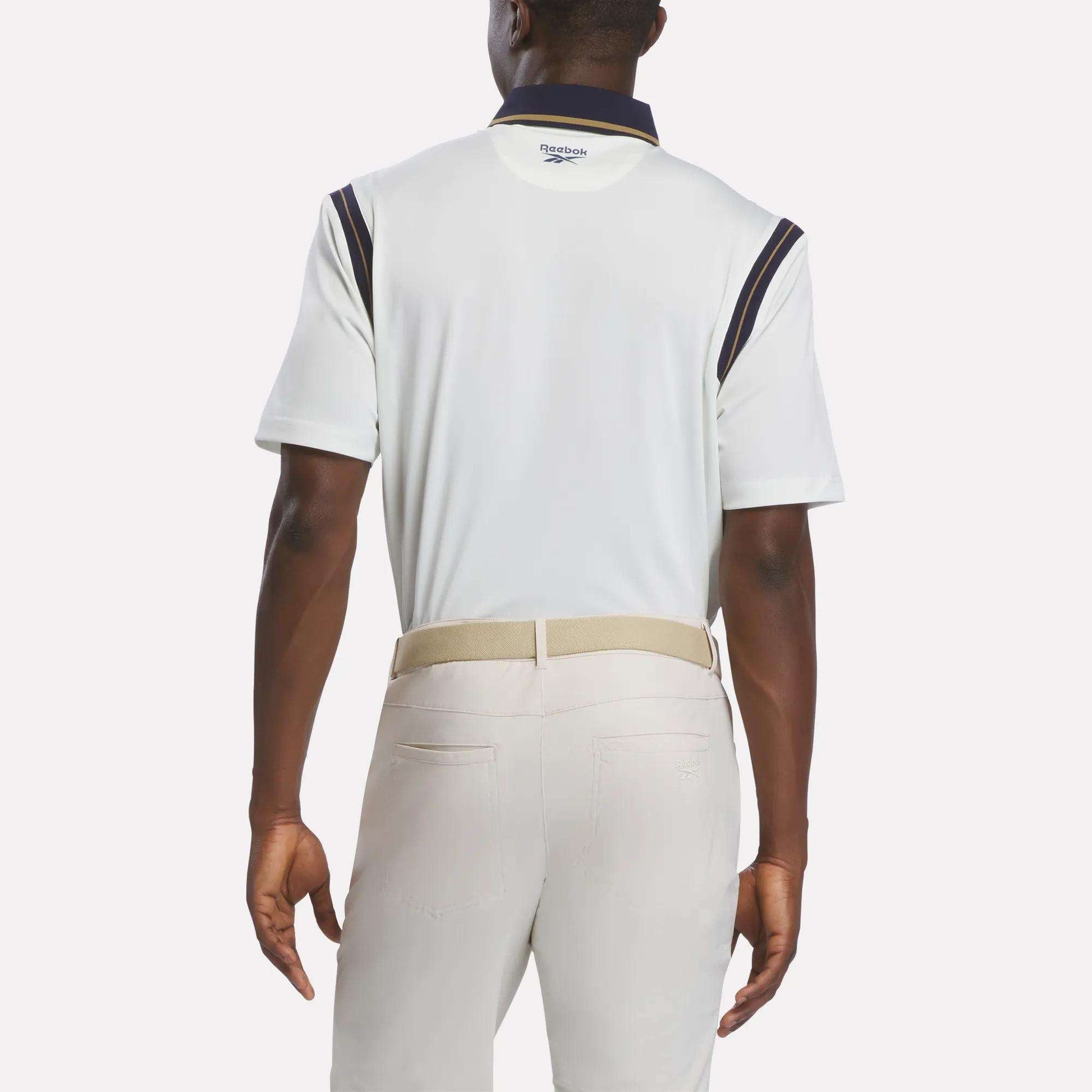 A man is shown from the back wearing a Men's Varsity Polo Shirt by Reebok, featuring RBK-DRY fabric with navy accents. He pairs it with light beige pants and a light-colored belt. The background is plain white.