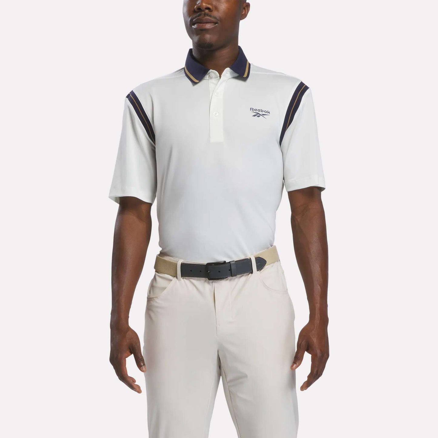 A man wearing a chalk-colored Men's Varsity Polo Shirt by Reebok, crafted from moisture-wicking RBK-DRY fabric and featuring navy accents, along with beige pants and a black belt, stands with his arms relaxed at his sides against a plain background.