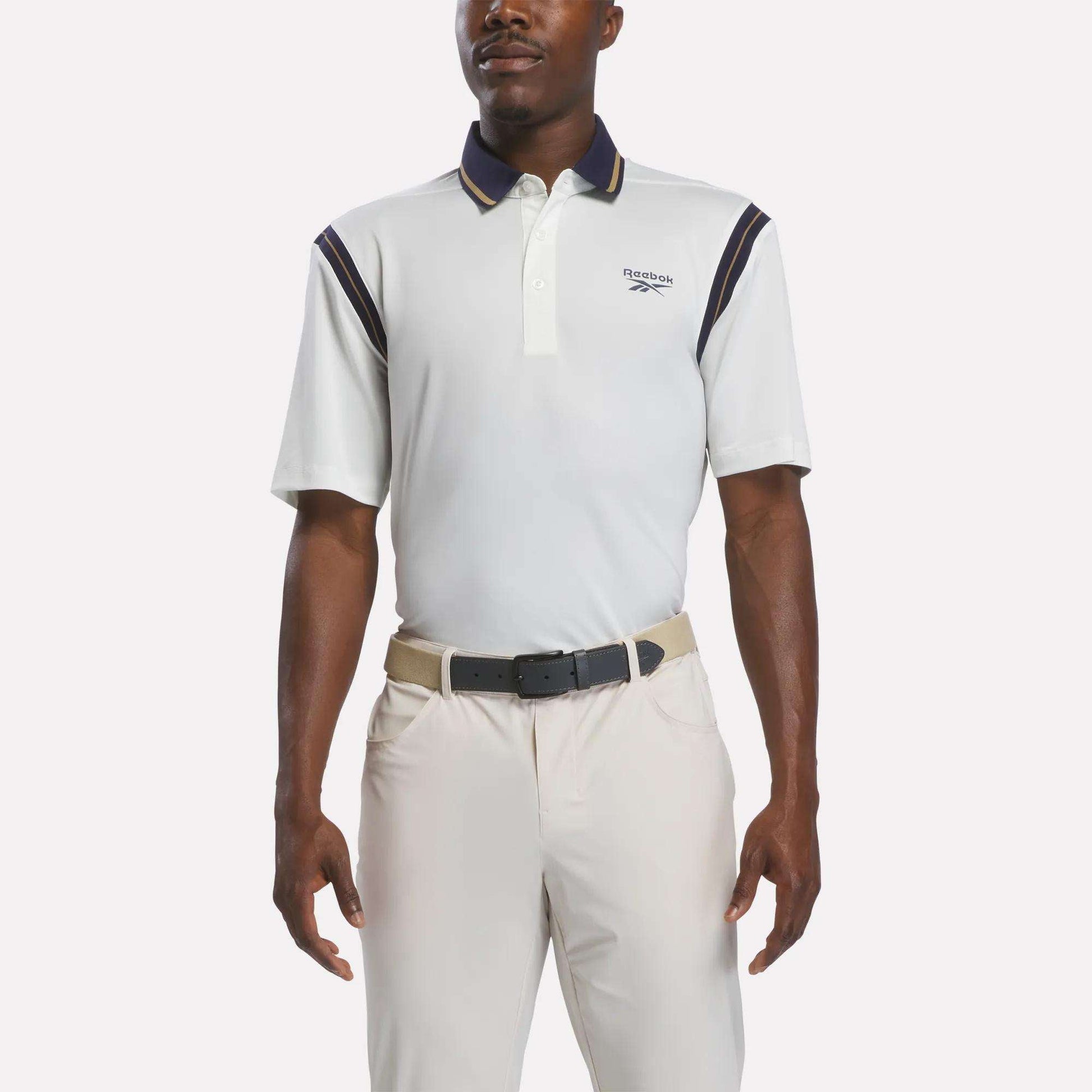 A man wearing a chalk-colored Men's Varsity Polo Shirt by Reebok, crafted from moisture-wicking RBK-DRY fabric and featuring navy accents, along with beige pants and a black belt, stands with his arms relaxed at his sides against a plain background.