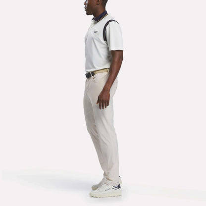 A man stands in profile wearing a crisp Reebok Men's Varsity Polo Shirt, paired with light beige pants and white sneakers. His hands are relaxed by his sides against a plain white background.
