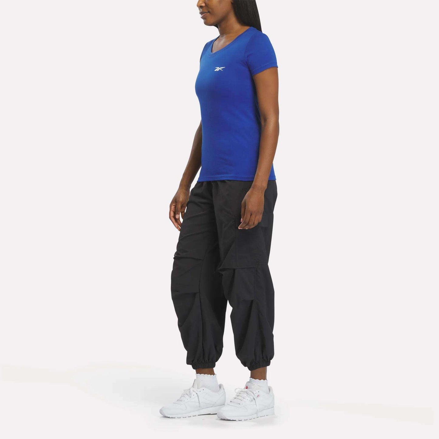 A woman faces left against a plain white background, wearing a fitted blue Reebok Women's T-Shirt, black cargo jogger pants, and white athletic sneakers.