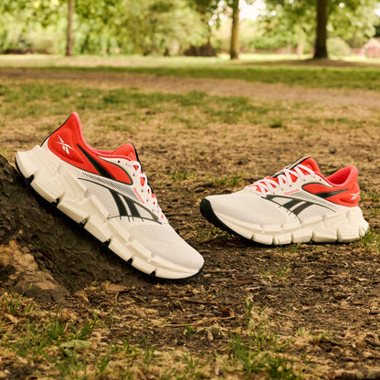 A pair of Reebok Men's FloatZig 2 Running Shoes in white with black and red accents rest on park grass, one leaning against a tree. Superfloat+ cushioning offers comfort, and the shoes stand out amid the greenery.