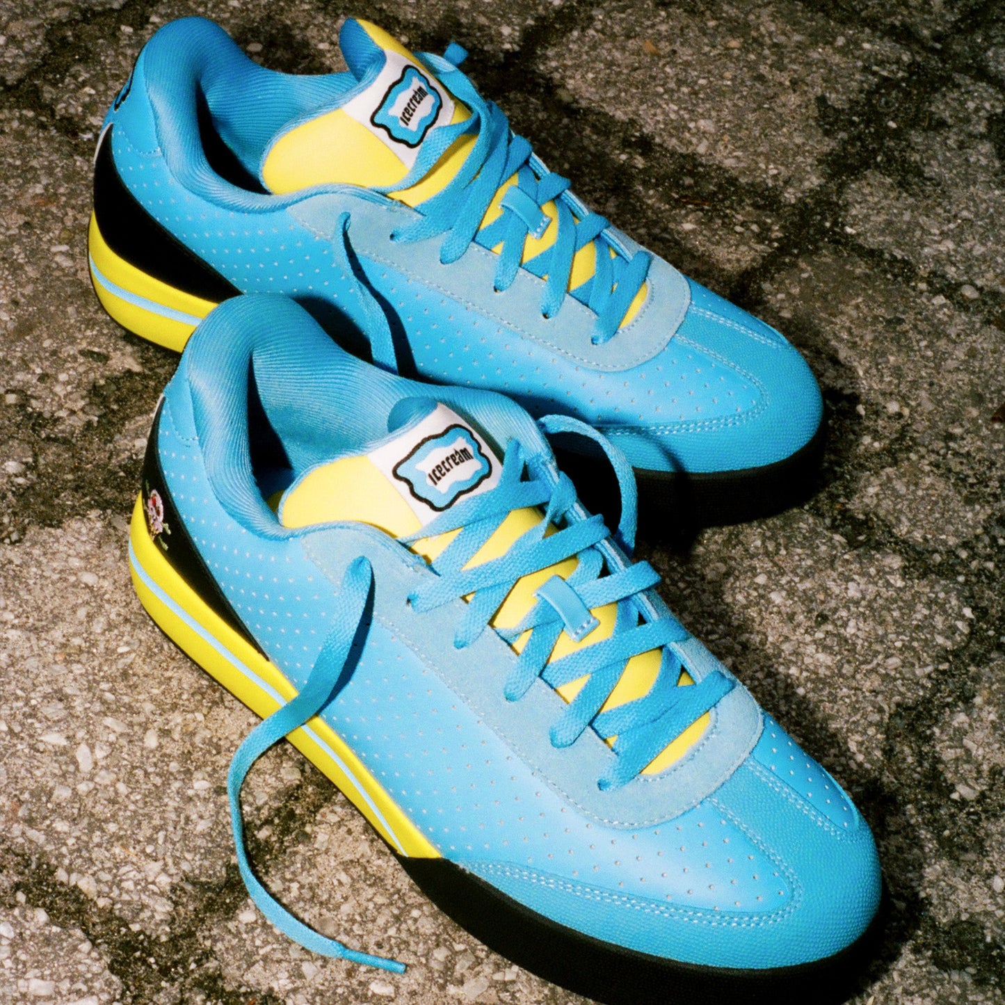A pair of Reebok x Icecream Board Flip Shoes with blue perforated suede uppers, yellow accents, blue laces, and black soles are placed on a stone pavement. Featuring a sporty design, the shoes display the Reebok logo on the tongue labels.