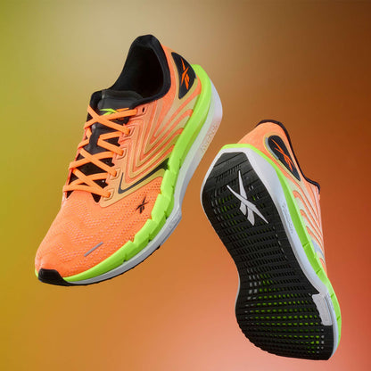Two vibrant orange Reebok Men's FloatZig Tread Running Shoes with neon green accents and cushioned midsoles are displayed on a gradient brown-orange backdrop. One shoe reveals the top and side; the other features the sole—ideal for treadmill workouts.
