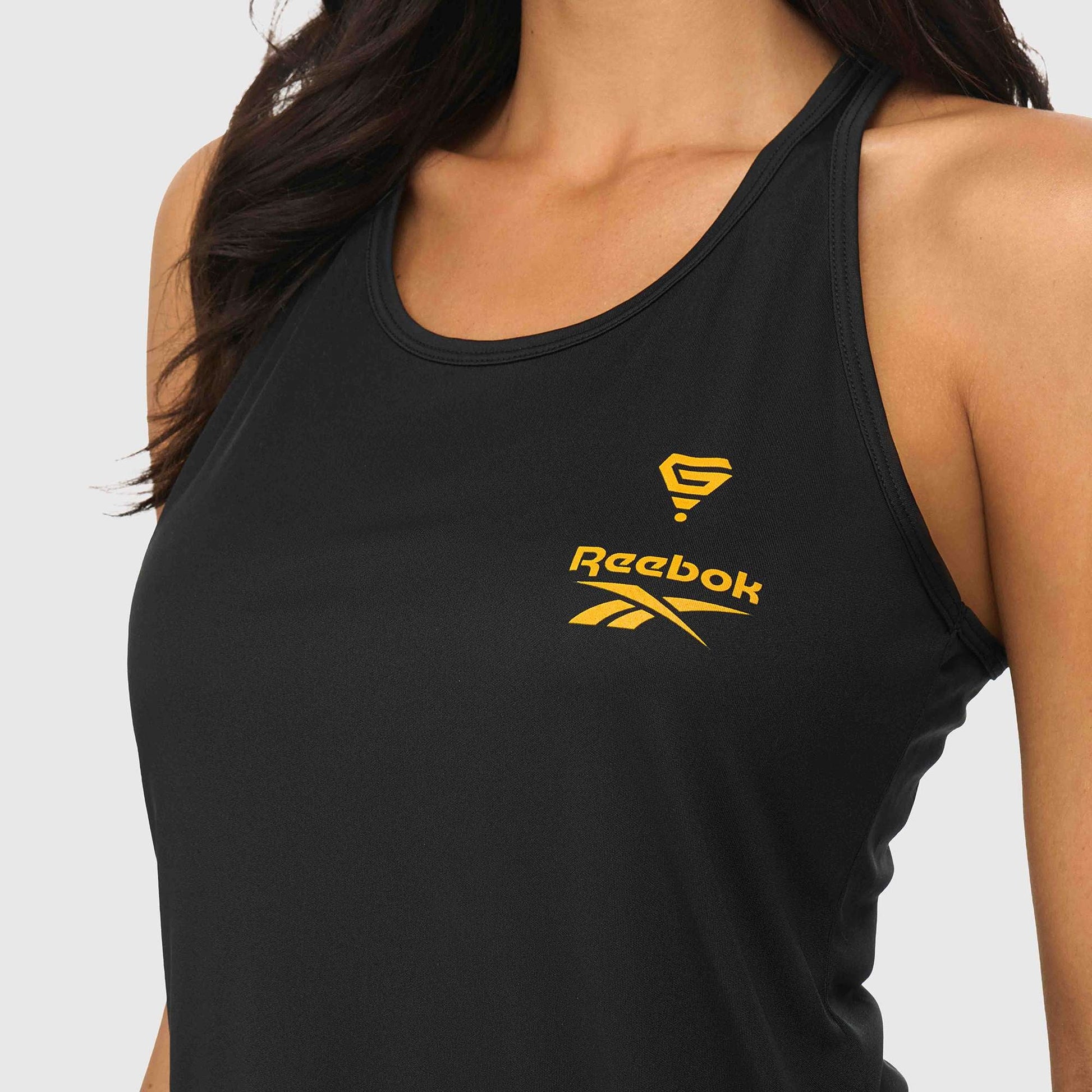 A woman wears the Reebok x GLDN PNT Women’s Tank Top, a sleeveless black Reebok athletic top with yellow logos on the front, covering her upper torso and shown against a plain light background.