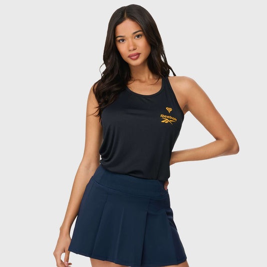 A woman with long dark hair wears a sleeveless black Reebok x GLDN PNT Women’s Tank Top with a yellow logo and a navy tennis skirt, standing with one hand on her hip against a plain light background.