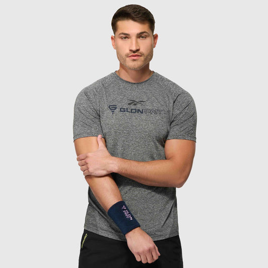 A man wearing a grey Reebok x GLDN PNT Tech Tee and black shorts stands against a plain background, holding his left arm with his right hand. He has a navy wrist support on his left wrist and a neutral expression.