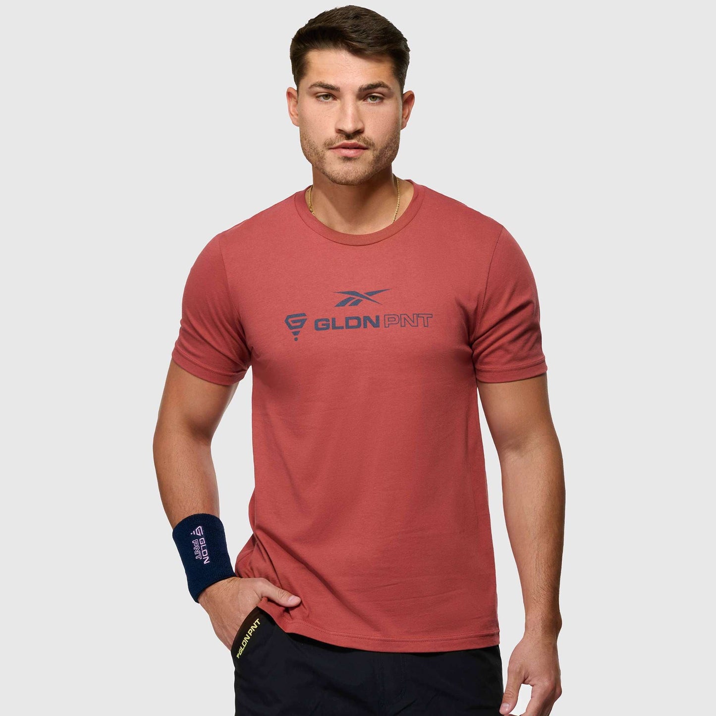 A man stands against a light gray background, wearing the Reebok x GLDN PNT Jersey Tee in salmon with a navy blue wristband and GLDN PNT logo, looking forward with a neutral expression and one hand in his pocket.