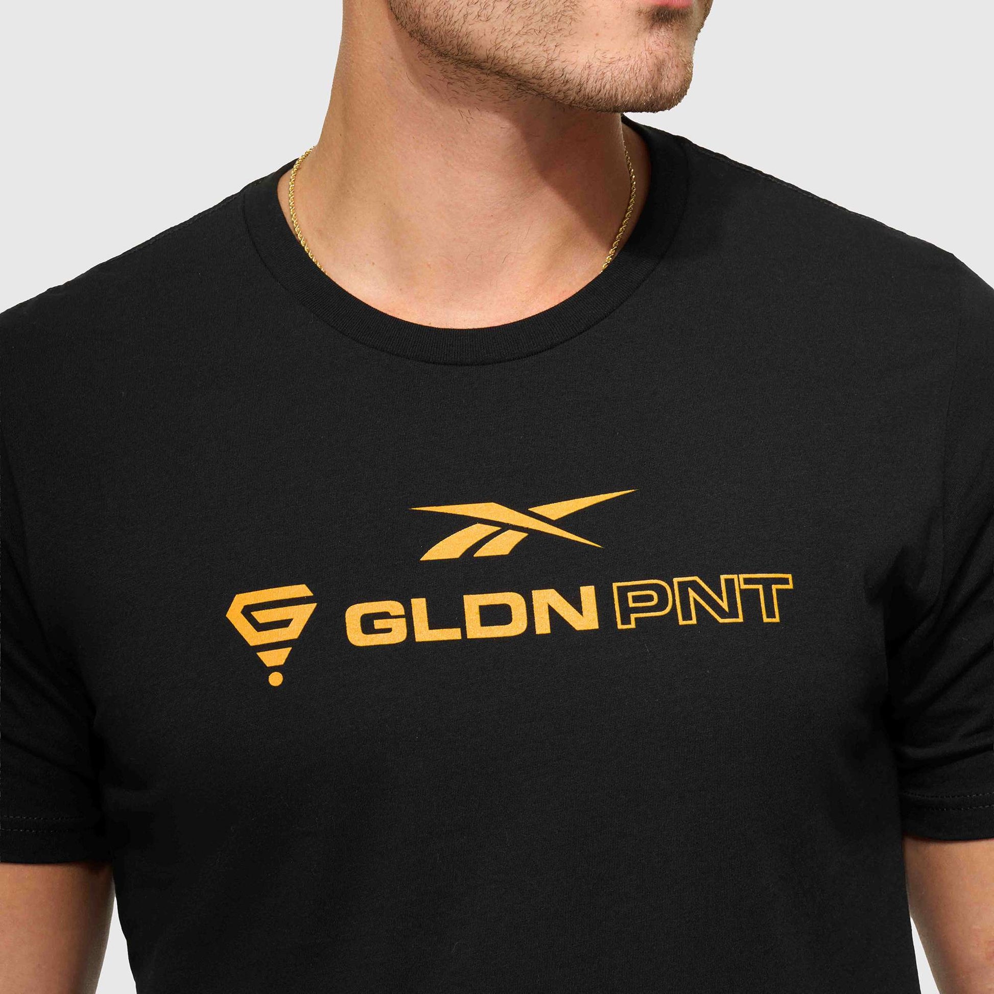 A person wears the Reebok x GLDN PNT Jersey Tee, a minimalist black unisex t-shirt by Reebok with gold graphics and 'GLDN PNT' text, made from premium cotton. Their face is partially visible as they look to the side.