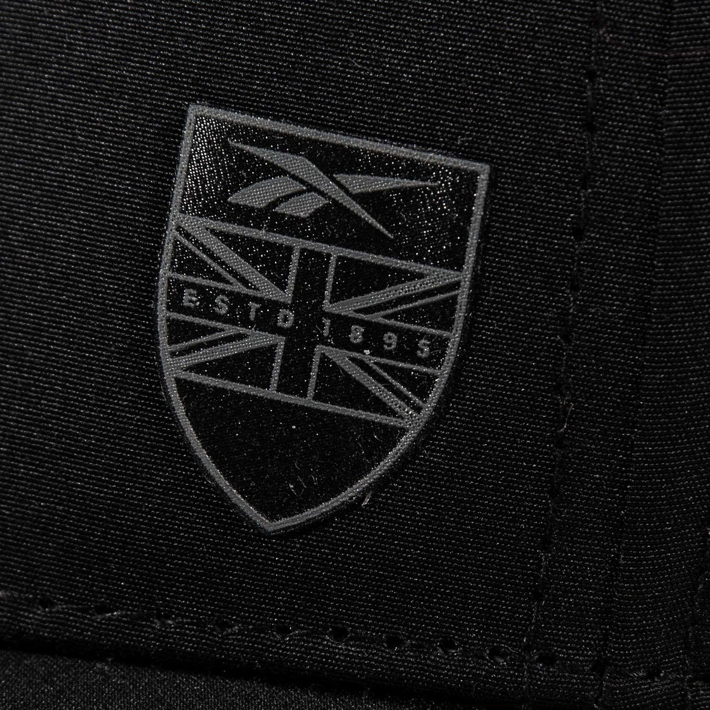 Close-up of black Chip Pro fabric by Reebok, showcasing a shield-shaped logo with the Union Jack pattern and gray "ESTD 1895" text.