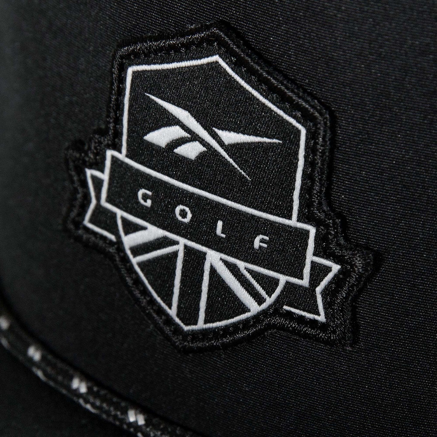 A close-up of the Madison Ii by Reebok shows a black patch with a white shield and stylized logo reading "GOLF" in bold, featured on a recycled polyester hat with an adjustable snapback.