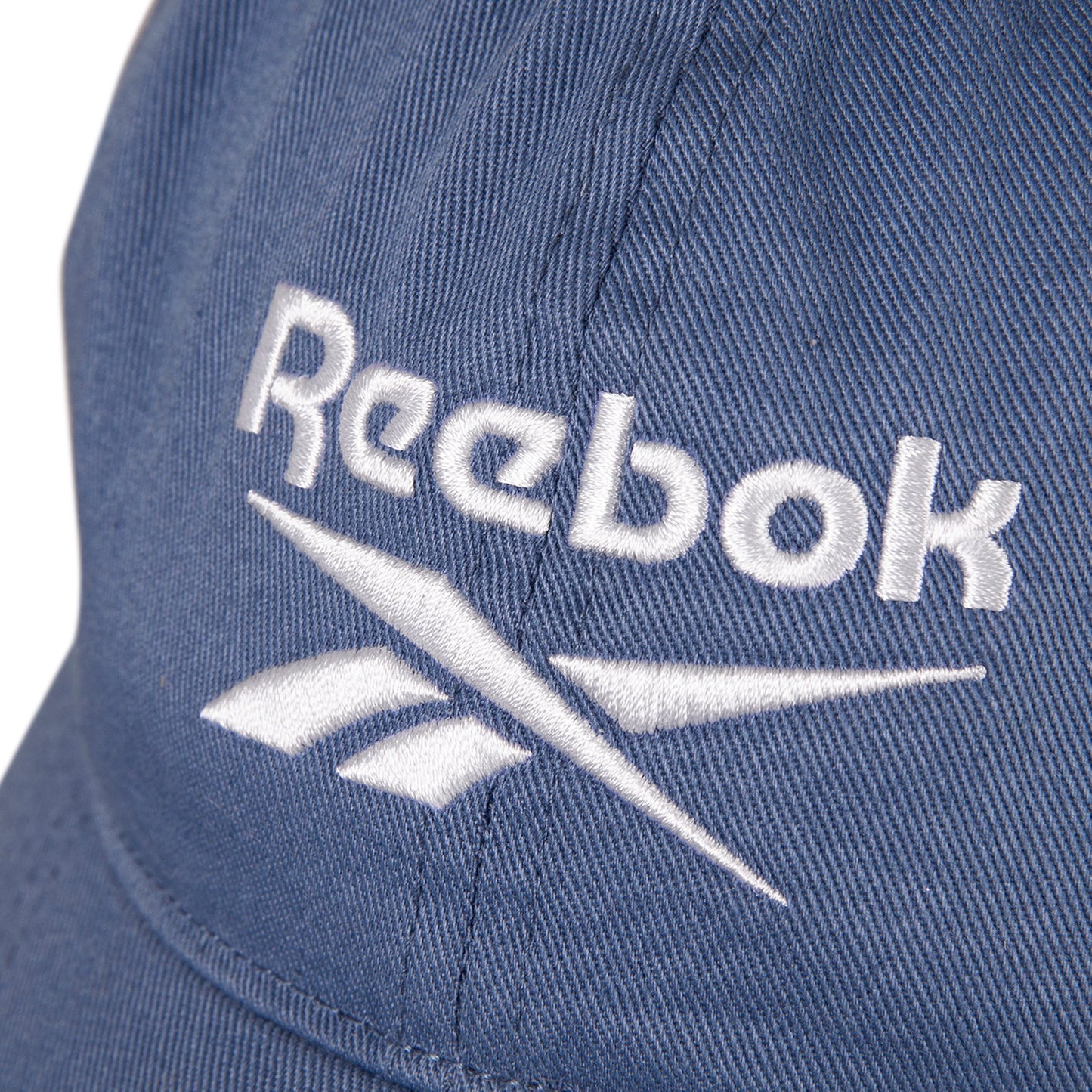 Close-up of the Reebok Logo Cap, made from cotton twill with a visible white embroidered Reebok logo and text. The cap features fabric texture details and an adjustable buckle.